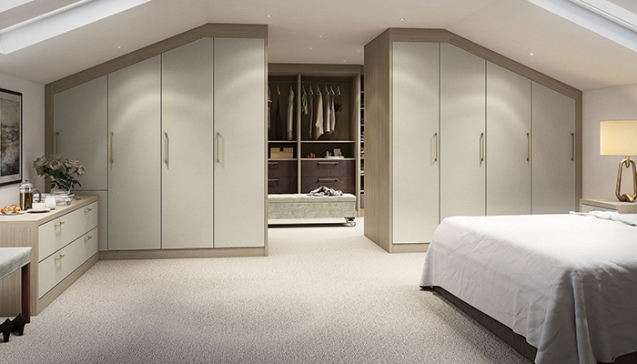 The double set of full-height, under-eaves fitted wardrobes, bedside tables and chest of drawers from Daval’s made-to-order, linen-effect Langham range are seen here in Silk Beige with brass bar handles. Additional storage is provided by the walk-in dressing room featuring low-level Mayfair drawers