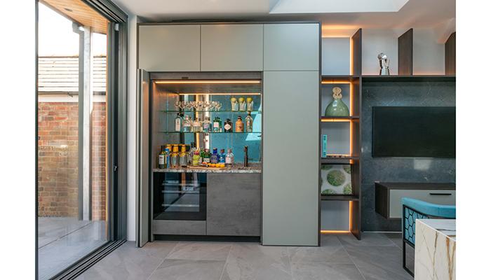 A cocktail bar cabinet next to the media area features a full-height bi-folding pocket door that slides into a side recess when in use and includes a push-to-open wine fridge by M’Quvée and small granite clad 1810 sink with Quooker tap in gun metal. Antique mirror splashback and glass shelves