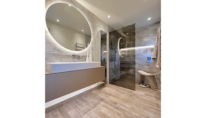 This spa-inspired master ensuite by The Tap End features a bronze-tinted shower screen and oversized backlit mirror, which is centrally positioned above the wide matt metallic vanity unit 
